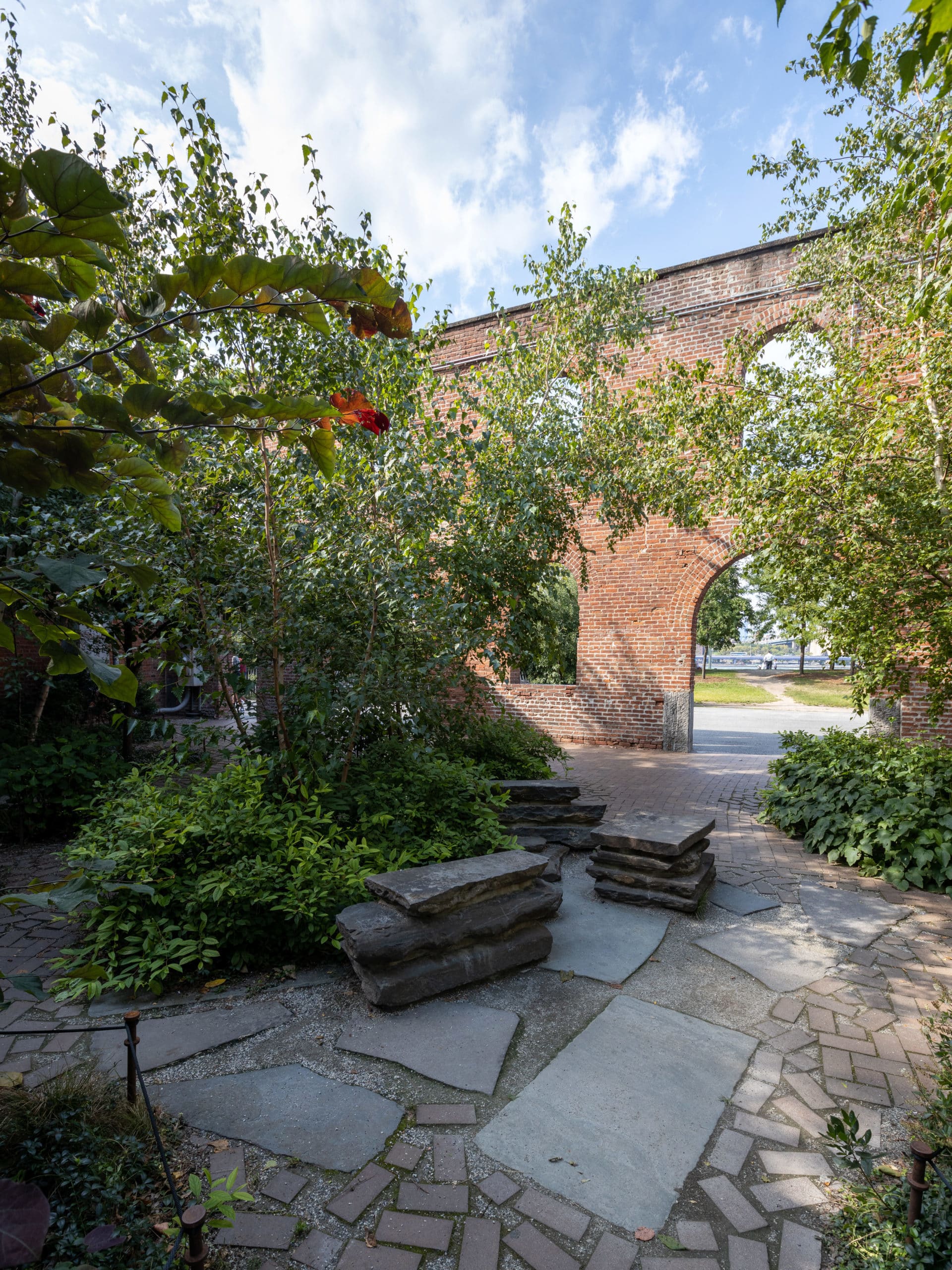 Pathway with stone benches in a garden with bushes and trees on a sunny day.