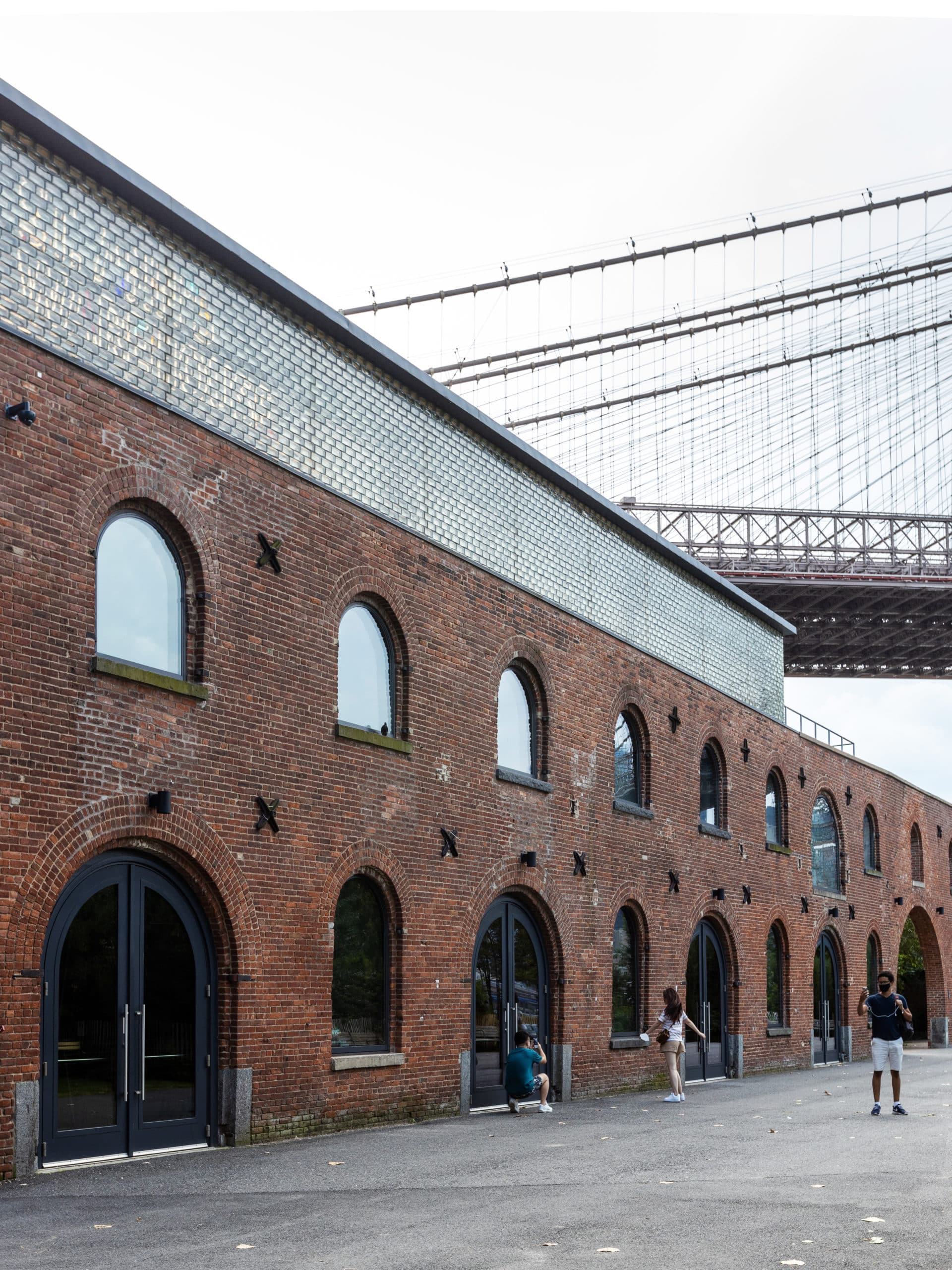 People posing outside St Ann's Warehouse on a cloudy day. The Brooklyn Bridge is seen overhead.