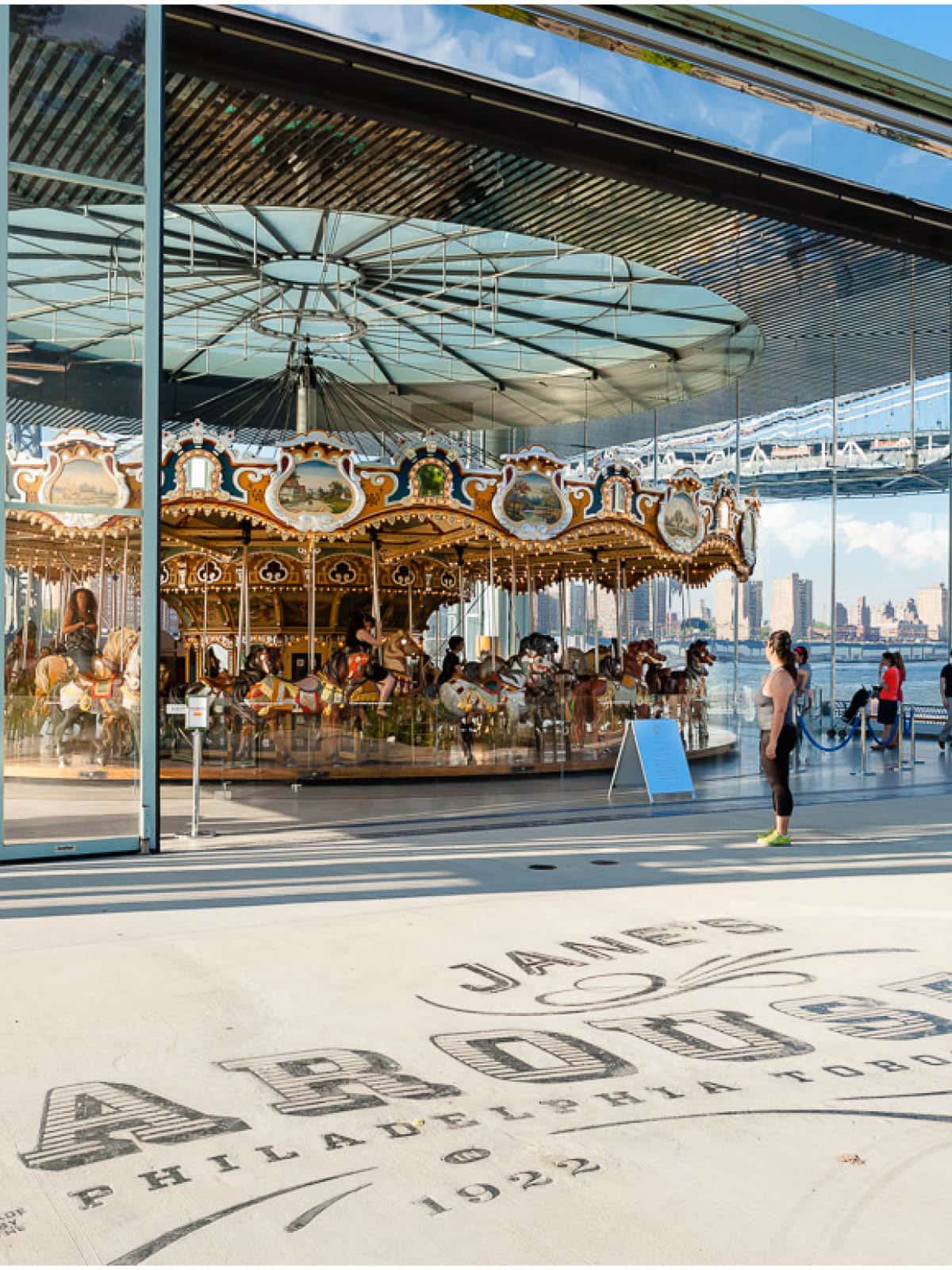 Jane's Carousel written on the sidewalk with the carousel in the background on a sunny day.