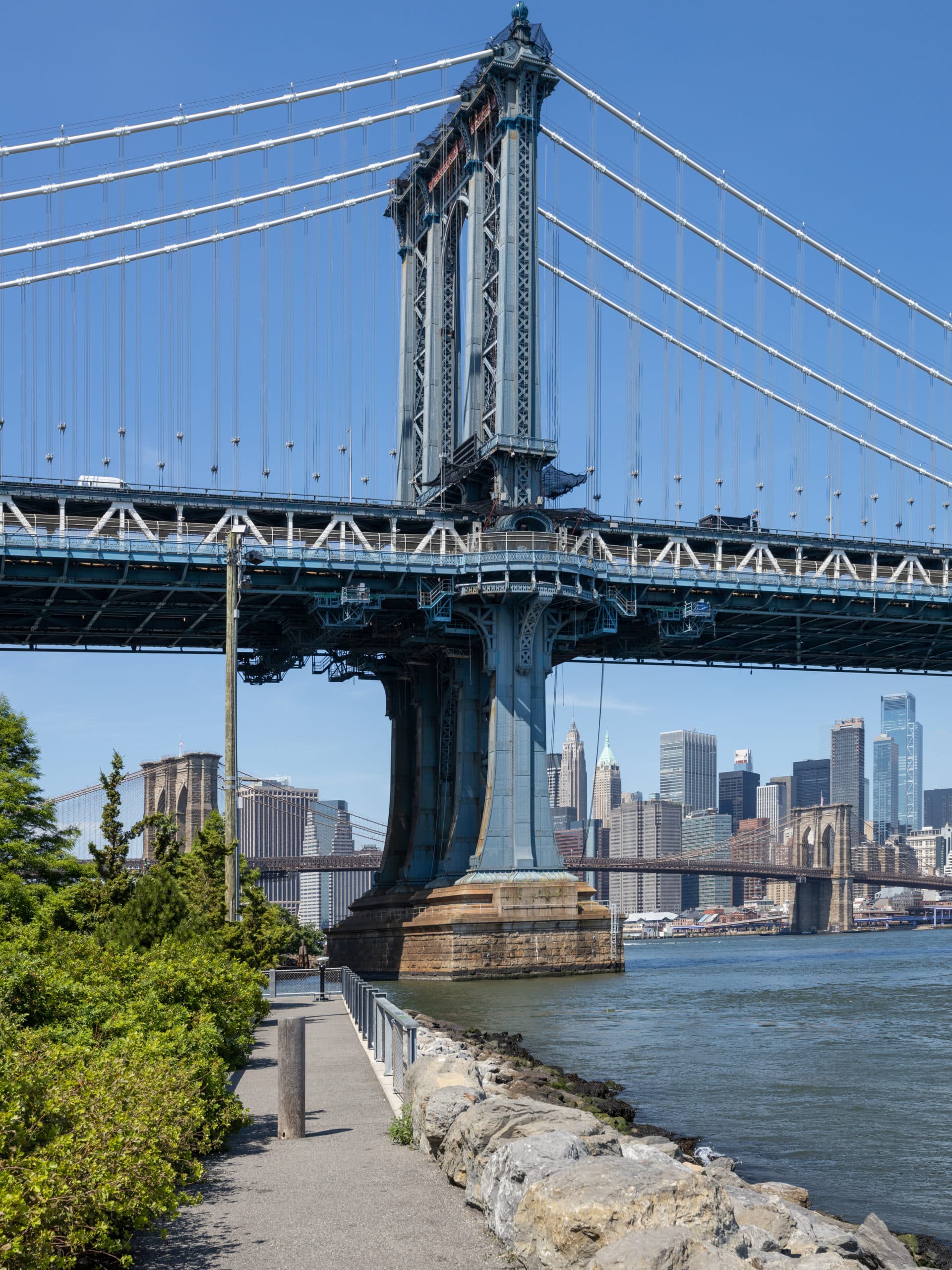 Pathway by the water with a view of the base of the Manhattan Bridge on a sunny day.