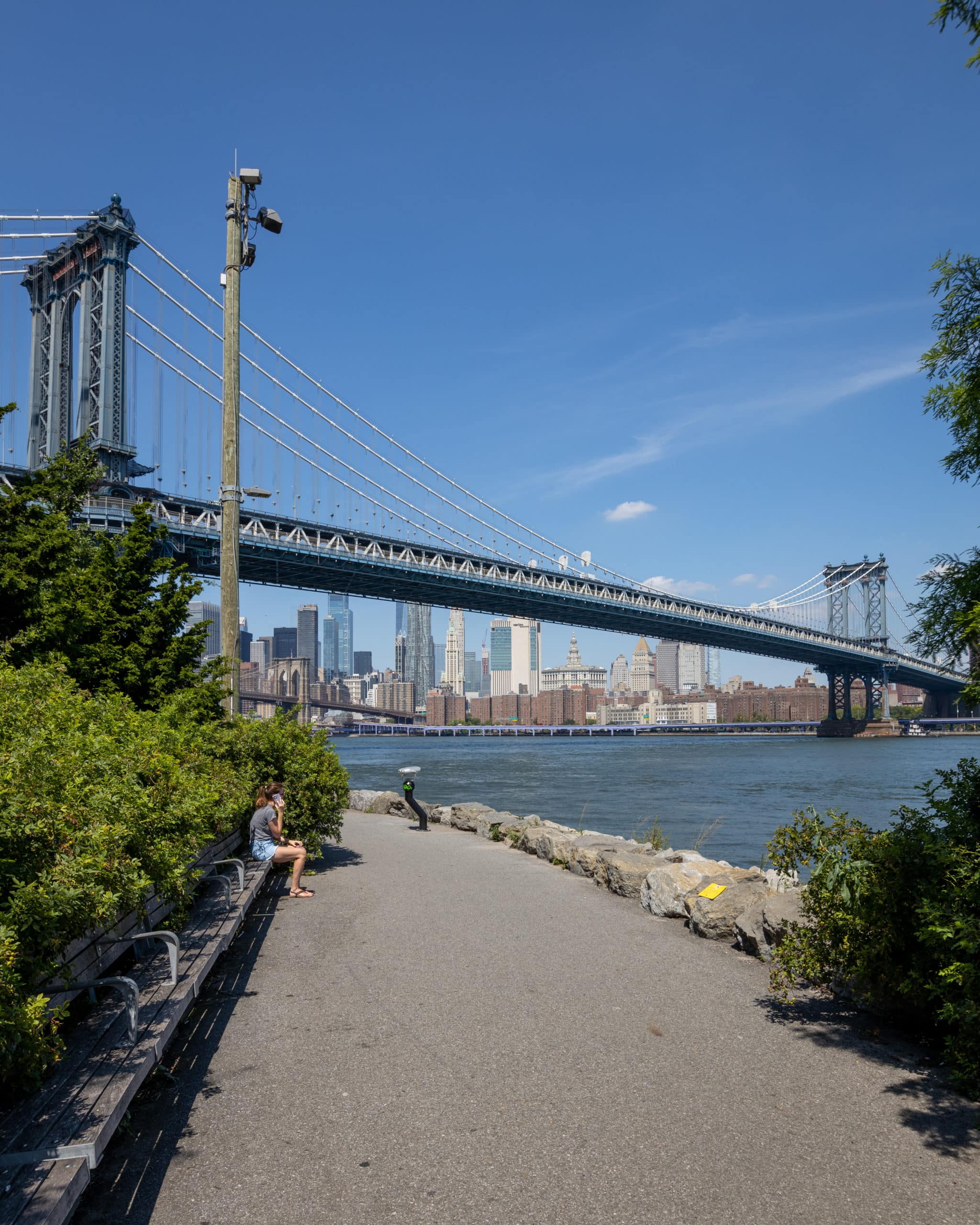 Woman sitting on benches by water on a sunny day. View of Manhattan Bridge and East River in distance.