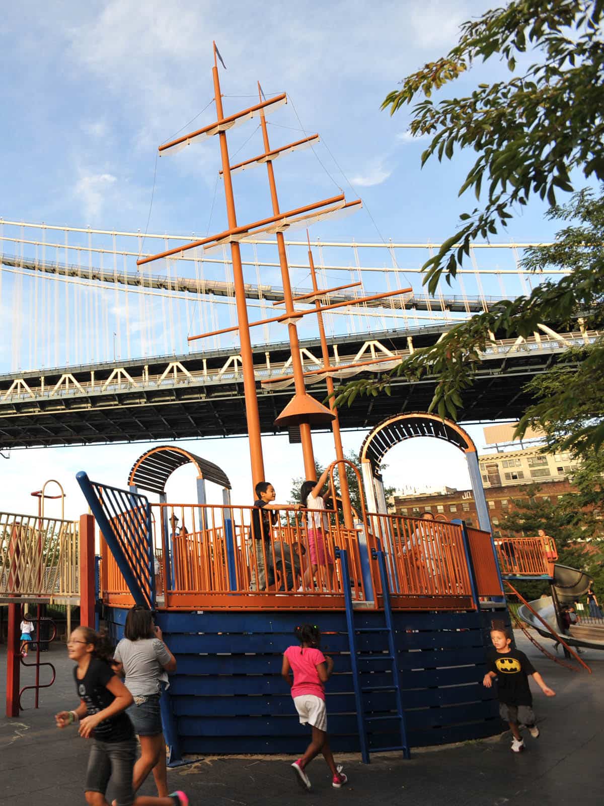 Children playing on a pirate ship themed playground structure on a sunny day. The Manhattan Bridge is seen overhead.