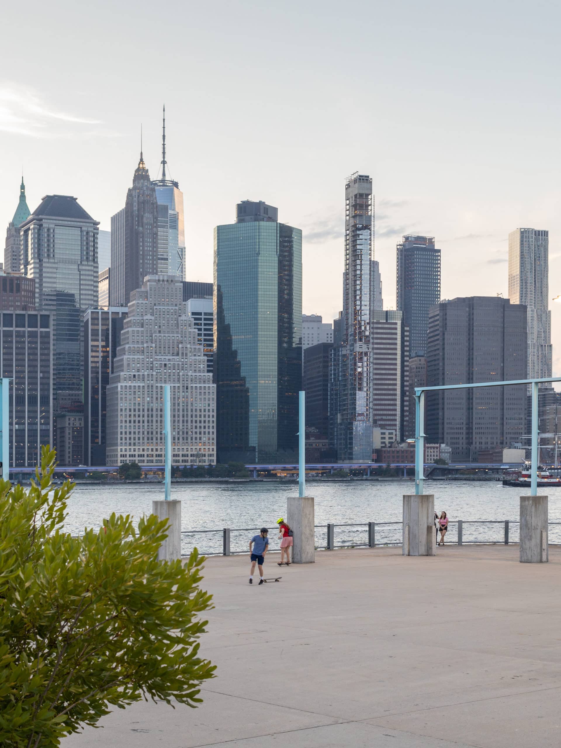 Kids skateboarding on Pier 3 Plaza with view of lower Manhattan in background at sunset.