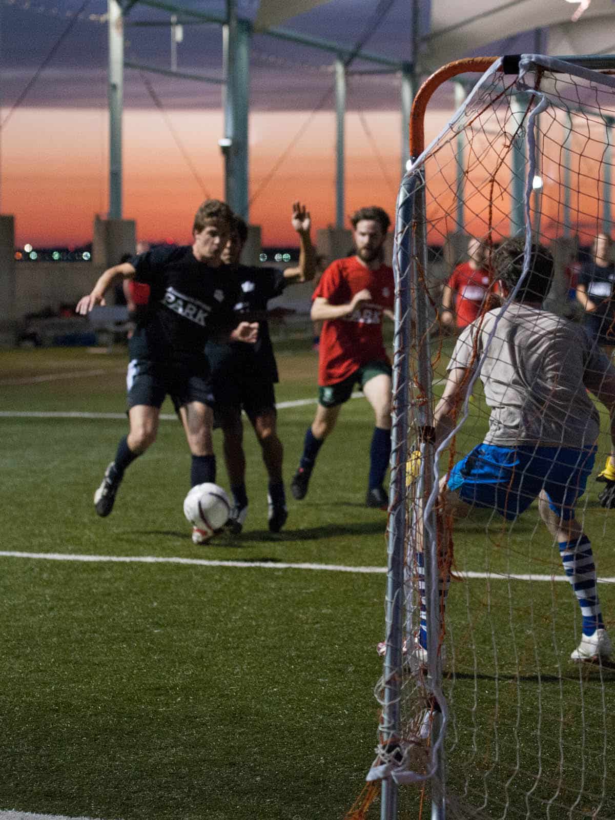 Men playing soccer by a soccer goal at sunset.