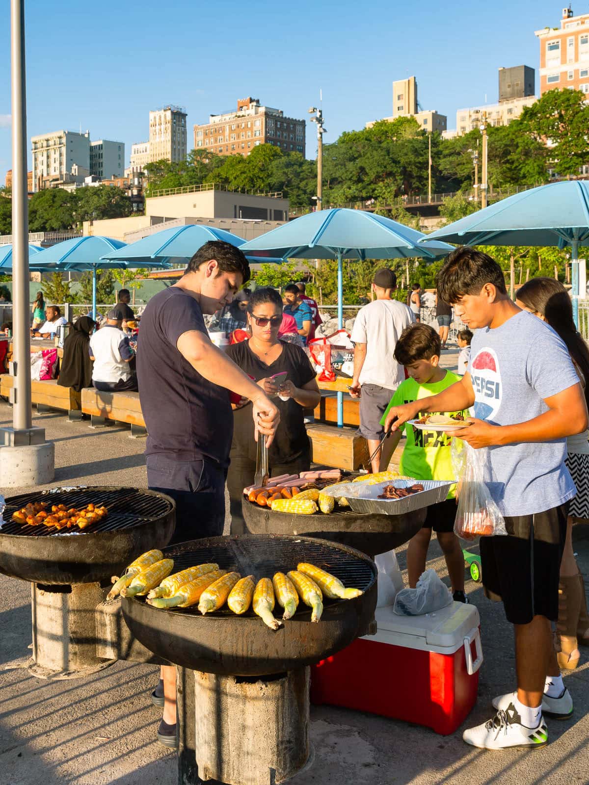 People around a grill with picnic benches in the background at sunset.