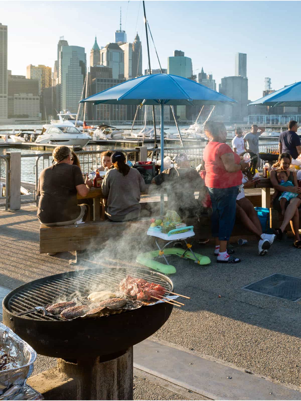 Food on the grill with people sitting behind at picnic tables at sunset.