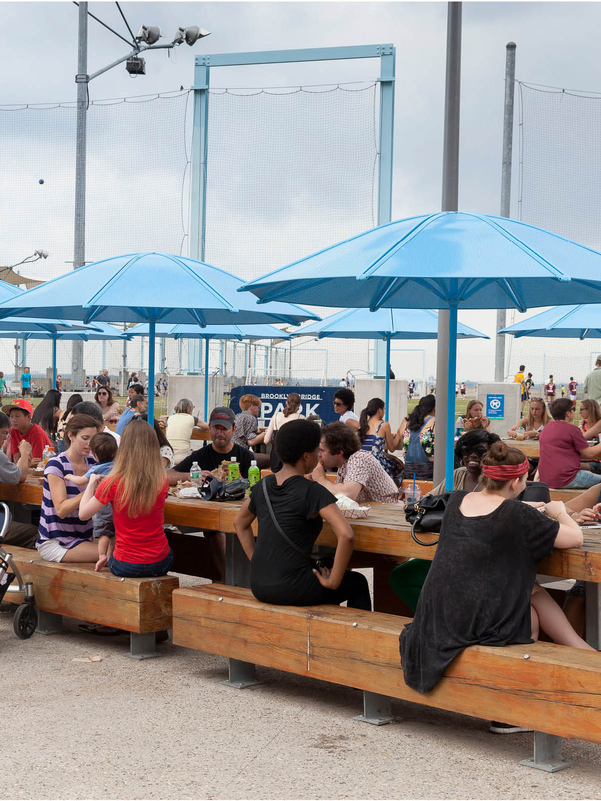 People eating at picnic tables with umbrellas on a cloudy day.