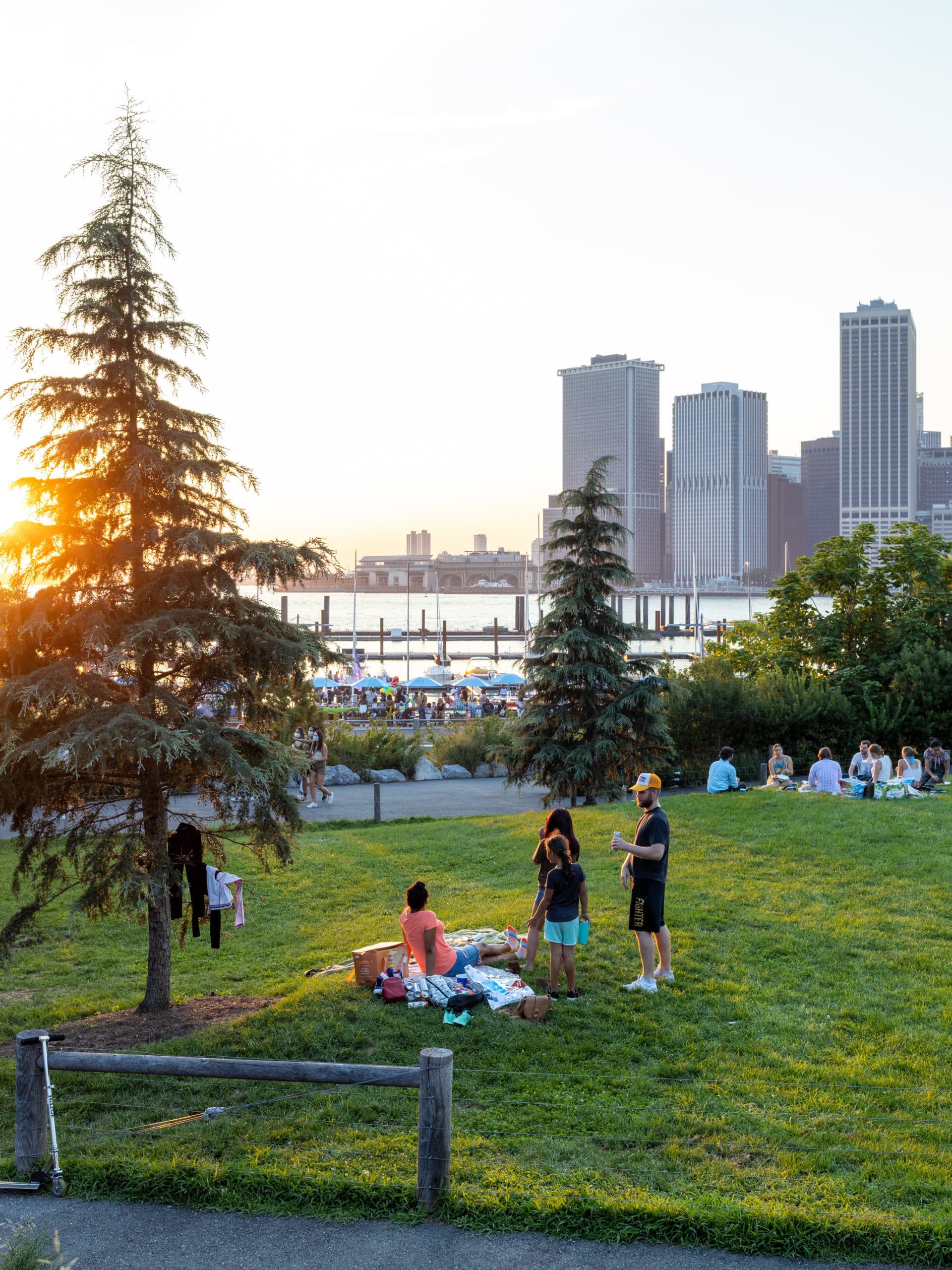 Family having picnic on Pier 5 Uplands Lawn at sunset. ONE°15 Brooklyn Marina and lower Manhattan seen in background.