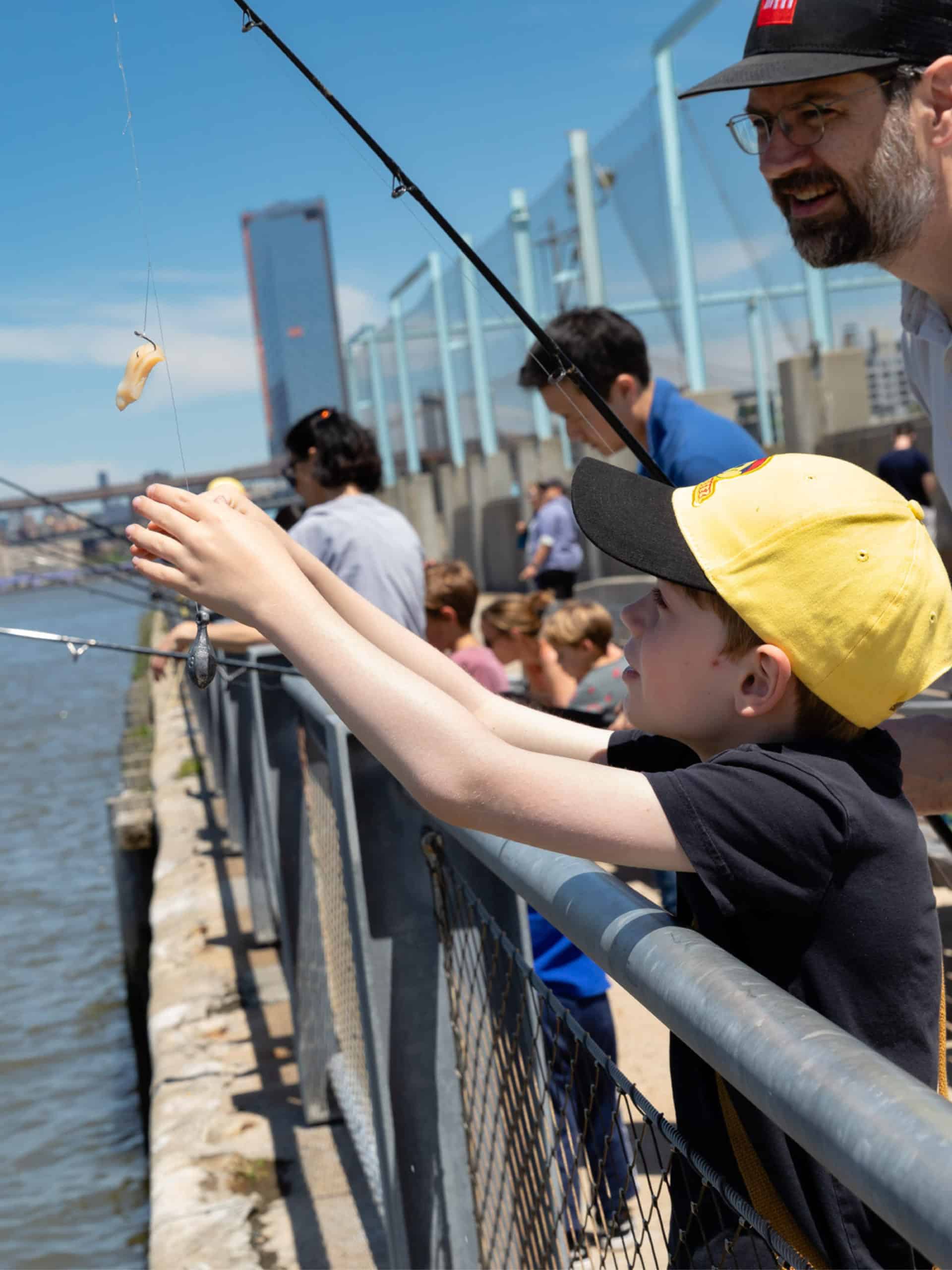 Close up of man and young boy with a fishing rod on a sunny. The boy is reaching for the hook. Other fishers seen in the background.