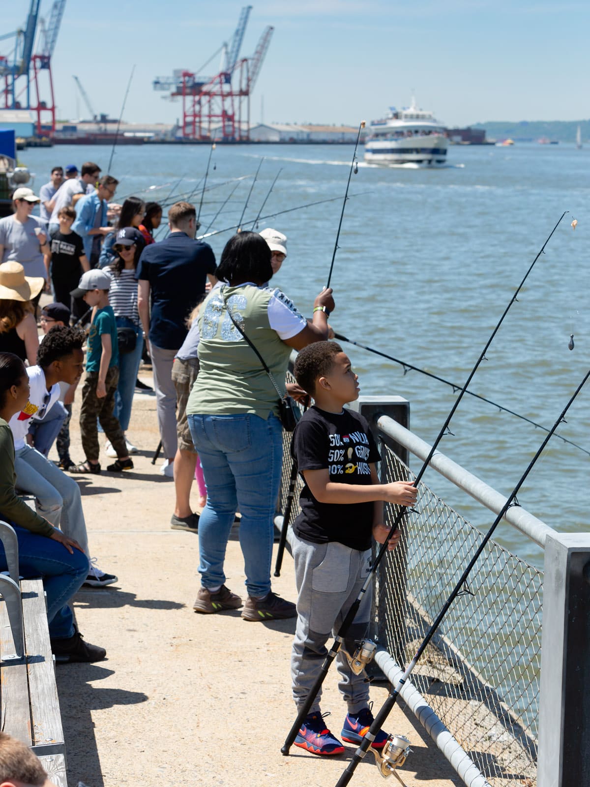 Group of people fishing along the promenade on a sunny day. Young boy in the front looks up at fishing rod. NYC ferry seen in the background.