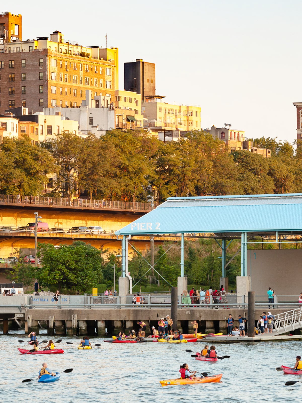 Groups of people kayaking by Pier 2 at sunset. Brooklyn Heights is seen in the background.