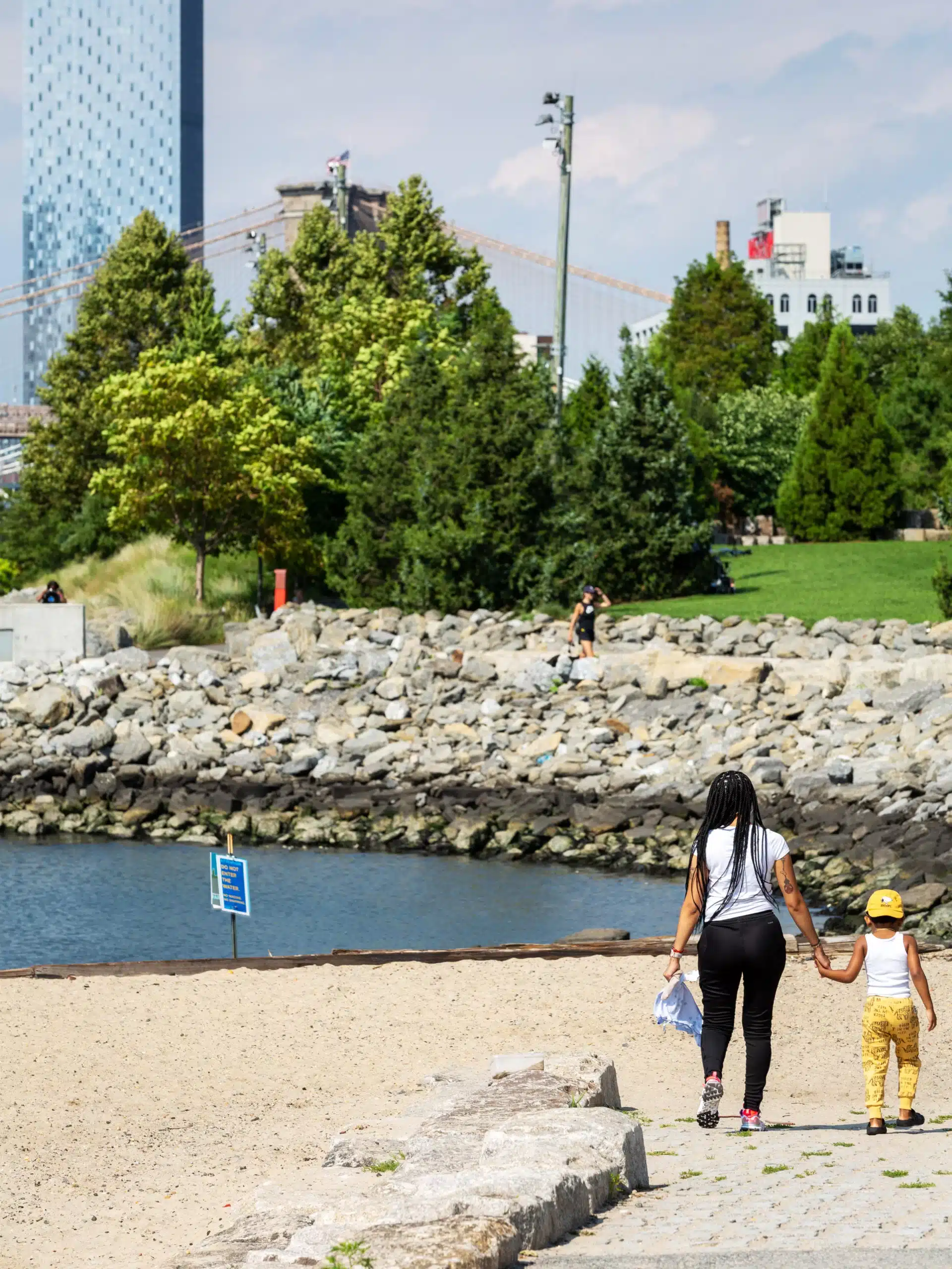 Woman and child holding hands walking down path to Pier 4 Beach on a sunny day. Pier 3 Greenway seen in the background.